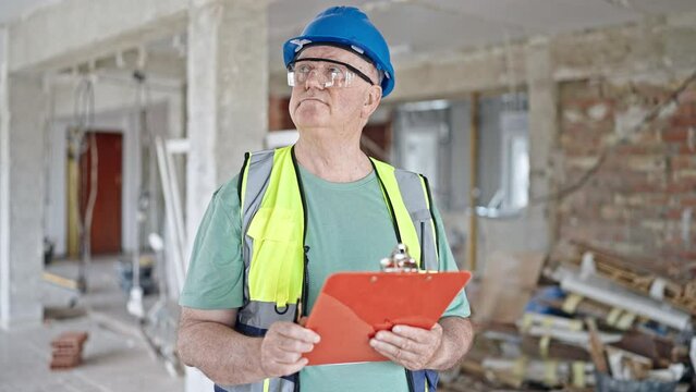 Middle age grey-haired man builder holding clipboard looking around at construction site