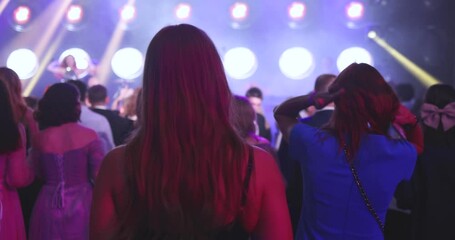 A crowded concert hall with scene stage lights, audience at a rock show performance with band performing music on a stage, with men and girls dancing on dance floor during a concert festival