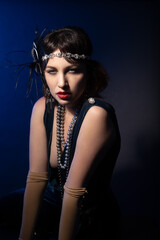 Studio portrait of a caucasian model wearing 1920s retro fashion. She is dressed as a flapper with a blue dress, headband with a feather and red lipstick. The background is blue. 