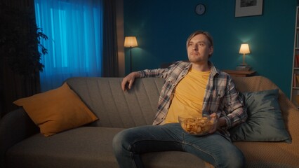 A man watches TV at home in the evening. A man is sitting on the couch with a plate of chips and...