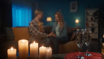 Glasses of champagne and a blurred loving couple in the background. Enamored man and women hold hands while sitting on the sofa in the living room. Romantic candlelit evening with champagne, date.