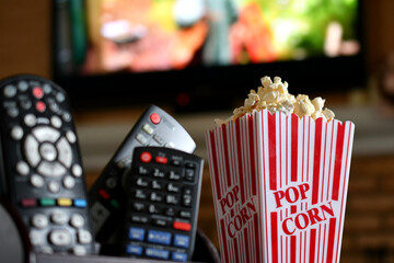 TV remotes and a container of popcorn with a blurred large screen television