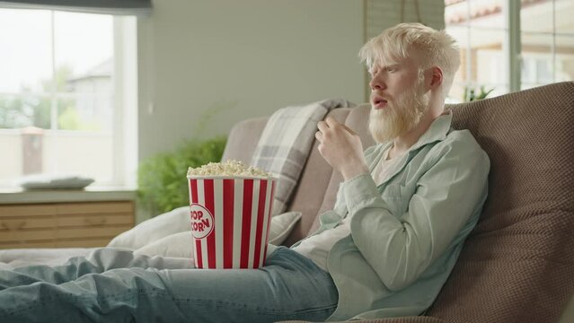 Excited bearded albino man sitting on sofa in living room, eating popcorn, watching TV. Albino man staring at screen, enjoying TV program. Leisure time. High quality 4k footage