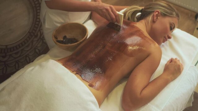 young blonde woman has beauty procedures at spa salon Slow motion. Beautician applying chocolate scrub on body parts with brush, medicine. Female patient lying on couch at wrapping treatment.