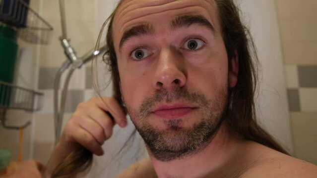 Close up of unshaved man try to comb his long dry untidy hair while standing shirtless in bathroom.