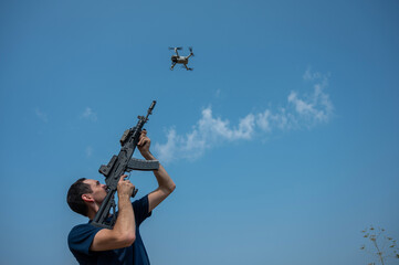 A man aims to shoot a rifle at a flying drone against a blue sky. 
