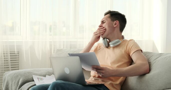 A tired Caucasian freelancer, working with documents and his laptop on the sofa. The exhaustion is evident in his demeanor, yet his determination to complete his tasks remains unwavering.