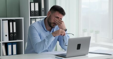 Caucasian businessman sits at his desk, struggling to keep his eyes open. He removes glasses, massages temples, and takes a deep breath in an effort to push through his exhaustion.