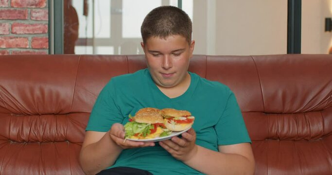 cheerful overweight boy whirling a plate full of burgers, choosing the tastiest of them, eating fast food with pleasure indoors Slow motion