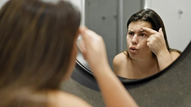 Young beautiful hispanic woman looking for pimples at the mirror in the bathroom