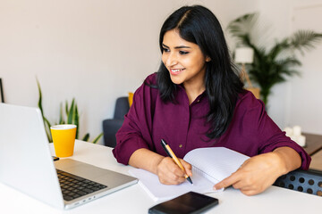 Young adult indian student woman taking notes while using laptop computer at home. Millennial ethnic female learning online listening virtual video call. Business and education concept. - obrazy, fototapety, plakaty