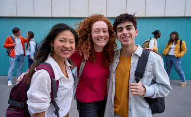 Group portrait of three smiling multiracial students hugging backpacks looking at camera standing outside college campus. Cheerful young people on break from classes. Generation z concept education. 
