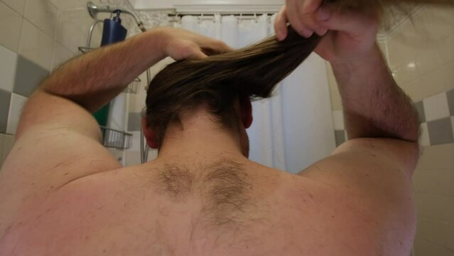 Close up of shirtless man ruffle and try to comb his long hair while standing in bathroom backwards to camera.