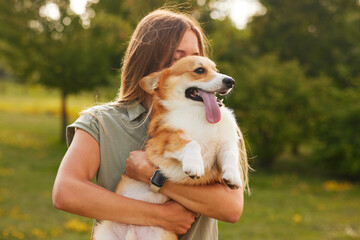 a young girl holds a cheerful and funny Welsh Corgi in her arms in a park in sunny weather, the concept of happy dogs