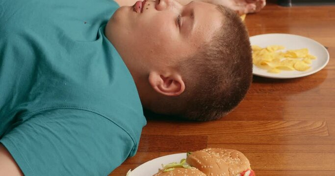 overweight plump teenager boy is full as he eats much oily fryed snacks fast food burgers chips Slow motion glutton with health problems cannot move. overeating, child is sleeping after breakfast