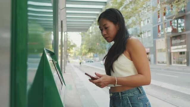 Young woman buys ticket at public transport stop using mobile phone