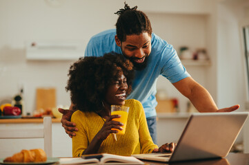 A happy african american couple is looking at the laptop and laughing at home together. - Powered by Adobe