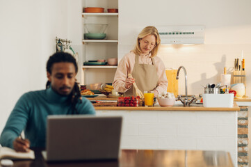 A blonde housewife is cooking in a kitchen while her interracial husband is working a freelance job on a laptop.
