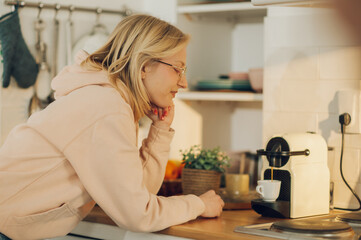 A sleepy woman is waiting for her morning coffee to be prepared while standing in a kitchen at home.