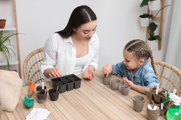 Mother and her daughter planting vegetable seeds into peat pots with soil at wooden table indoors