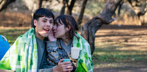 Two friends whispering in the cold, drinking tea covered with a blanket. Campfire at the campsite