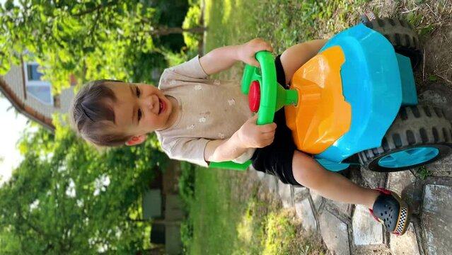 Caucasian baby boy riding cheerfully his toy car. Joyful time spending outdoors in summer. Vertical screen.