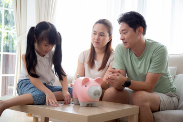 Obraz na płótnie Canvas Happy asian family father and mother and daughter sitting on sofa planning finance with saving while dropping coins in piggybank in room, mom and dad and kid deposit and budget, lifestyles concept.