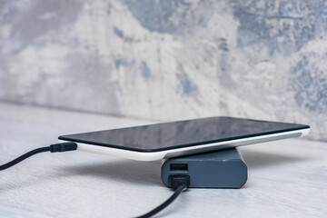 Tablet charging with power bank on a gray wooden table. Portable charger for charging devices.