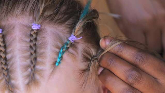The hands of an African woman are braiding the braids of a little girl close up view.