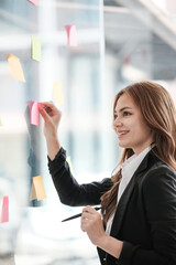 Beautiful female employee write down on colorful sticky notes manage list, concentrated biracial woman work on startup brainstorm collaborate plan on stickers on glass wall.