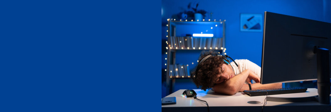 Arabic exhausted young man in headphones sleeping while playing video game on computer at home in blue neon light. Banner.