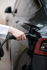 Young woman traveling by electric car having stop at charging station standing plugging charger close-up.