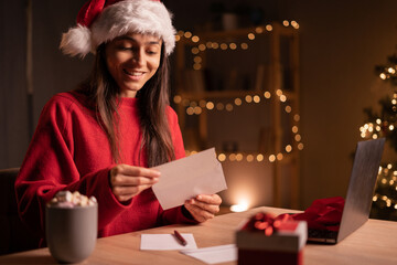 Young smiling woman sitting at desk at home and holding a envelope with christmas card. Christmas wishes, holidays and celebrations concept.
