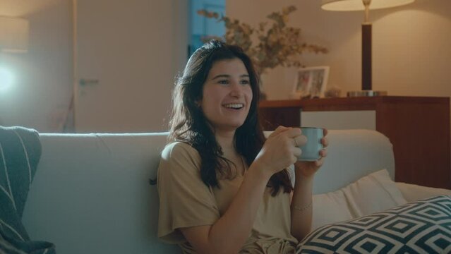 Young joyous woman sitting on sofa, watching interesting TV show, smiling and holding tea mug in the living room at home