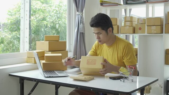 Asian young man selling online at home with box and laptop taking orders from customers, concept with SME business