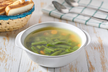 Delicious vegetable soup with green beans, potato, and carrot on a white wooden table.