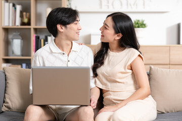 Fototapeta premium Asian young couple sit on couch or sofa looking at Computer Laptop smile and laughing together. Cheerful lover surfing internet and social media spending time in holiday at cozy home. Valentine Day