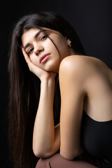 Classic studio portrait of a young brunette dressed in a black top, who is sitting on a chair against a black background.