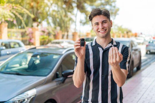 Young handsome man holding car keys at outdoors inviting to come with hand. Happy that you came