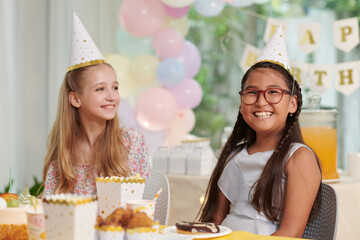 Portrait of cheerful preteen girl sitting at table at birthday party of her best friend
