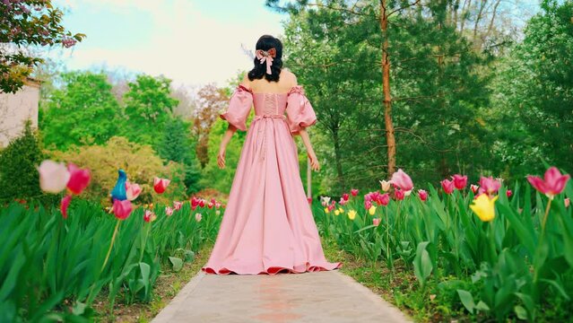 Fantasy girl princess walks in blooming spring garden flowers tulips tree green grass alley. Woman queen in long royal pink dress with train puffed sleeves vintage old style. lady back rear view. 4k