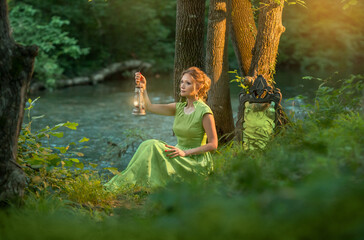 girl is sitting in the forest in a green long dress with old kerosene lamp in her hands