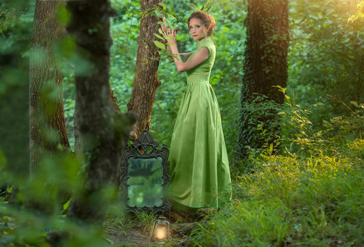 girl in a green long dress stands in the forest with an antique mirror