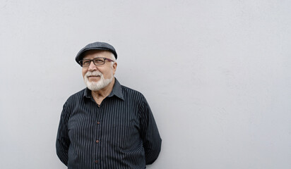 An elderly smiling man in glasses and a cap stands against a light wall background, hands behind his back, horizontal format