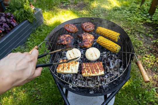 Hand mit Grillzange vor dem Holzkohlegrill mit K&auml;se Wurst und Fleisch belegt