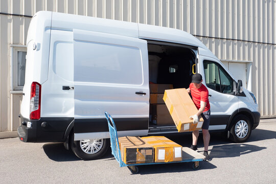 Courier unloads minivan. Man picks up cardboard box. Guy near courier car and cart. Process of unloading minivan. Courier in shorts and cap holds parcel. Working man near warehouse building