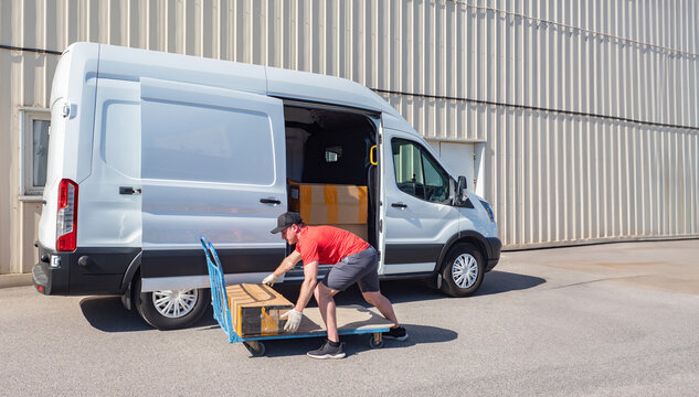 Courier unloads minivan. Delivery man picks up cardboard box. Guy loads parcels into courier car. Man near cargo cart. Courier with minivan near production building. Delivery service worker