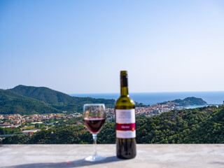a bottle of red wine and a glass against the backdrop of the sea and the town among the mountains