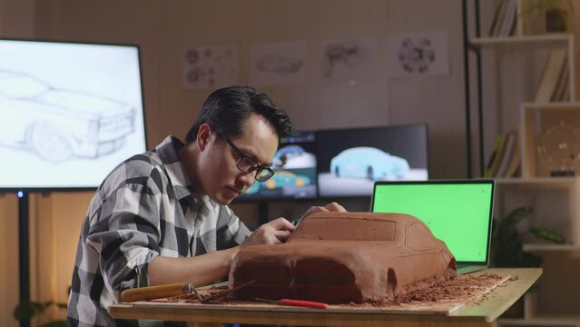 Asian Man Automotive Designer With Green Screen Laptop Using Rake Or Wire To Smooth Out The Surface And Create Details In The Sculpture Of Car Clay In The Studio
