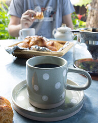 A cup of black coffee with it pot and porcelain on table while man eating French croissant in background with garden scenery 